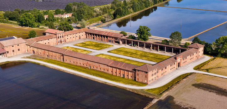 Aerial view of Tenuta Colombara a historic brick farmstead with a large courtyard surrounded by flooded rice paddies and green trees in Livorno Ferraris Piemonte Italy.
