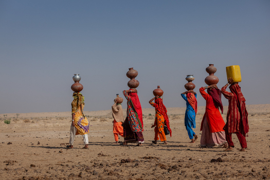 Yazman Punjab Pakistan 19 March 2010 View of women in traditional colorful clothing walking across the arid Thar desert landscape while carrying water pots on their heads near Chanan Pir.
