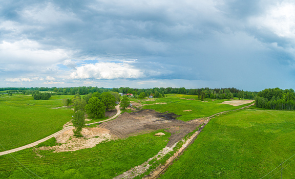 Aerial view of a rural landscape featuring lush green fields a dirt path winding through trees and a patch of dark soil under a dramatic cloudy sky in Latvia. Aerial view of a rural landscape featuring lush green fields a dirt path winding through trees and a patch of dark soil under a dramatic cloudy sky in Latvia.