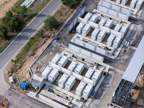 Aerial view of a battery energy storage system facility with rows of white containers concrete perimeter walls and adjacent road infrastructure in Poland.