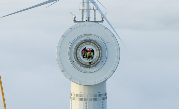Netherlands 26 November 2025 Aerial view of two technicians in high visibility gear and red helmets sitting inside the circular hub of a large wind turbine against a pale sky. Netherlands 26 November 2025 Aerial view of two technicians in high visibility gear and red helmets sitting inside the circular hub of a large wind turbine against a pale sky.