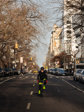 New York United States 13 April 2015 View of a cyclist in black gear and neon yellow shoes riding along a city street lined with bare trees and parked cars.