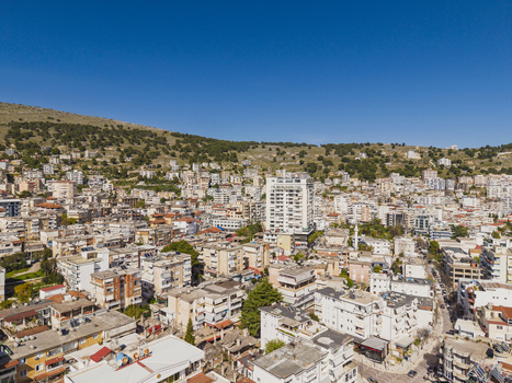 Aerial view of Saranda a coastal city in Albania featuring dense residential buildings a prominent white high rise and green hills under a clear blue sky. Saranda Vlore County Albania. Aerial view of Saranda a coastal city in Albania featuring dense residential buildings a prominent white high rise and green hills under a clear blue sky. Saranda Vlore County Albania.