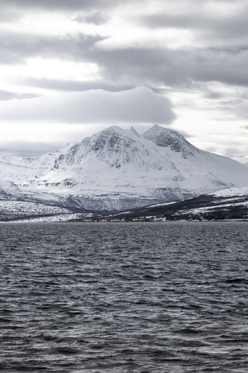 View of snow covered mountains rising above the dark waters of a fjord under a cloudy sky Tromso Troms Norway.