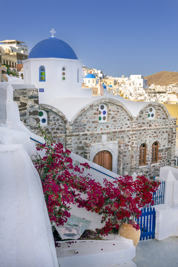 View of a traditional Greek Orthodox church with a blue dome and stone walls adorned with vibrant pink bougainvillea flowers under a clear blue sky in Lefkada Greece.