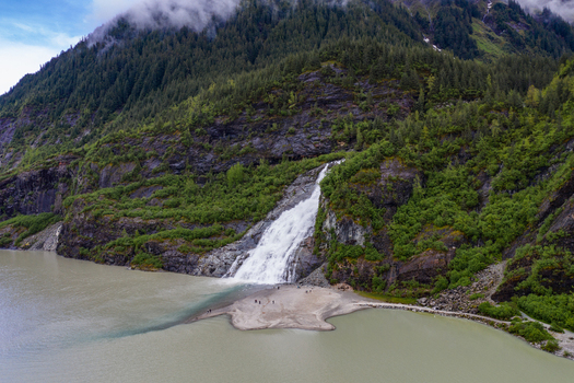 Aerial view of Nugget Falls cascading down a rocky cliff into Mendenhall Lake surrounded by lush green forest and misty mountains in Juneau Alaska United States. Aerial view of Nugget Falls cascading down a rocky cliff into Mendenhall Lake surrounded by lush green forest and misty mountains in Juneau Alaska United States.
