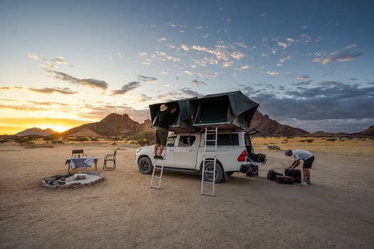 Spitzkoppe Namibia 12 March 2026 View of a camping setup under the vast sky with golden sunset hues painting the rugged landscape contrasting with the dark silhouettes of distant mountains.