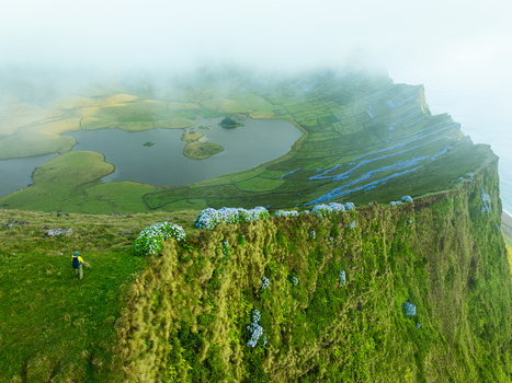 Aerial view of the Caldeirao volcanic crater lake on Corvo Island featuring lush green slopes blooming hydrangeas and a hiker on the cliff edge in Vila do Corvo Azores Portugal.