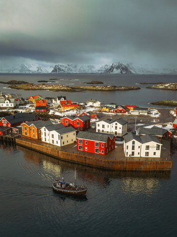 Aerial view of a fishing boat sailing into the harbor of a vibrant coastal village with colorful houses against the backdrop of snow capped mountains Henningsv&aelig;r Nordland Norway.