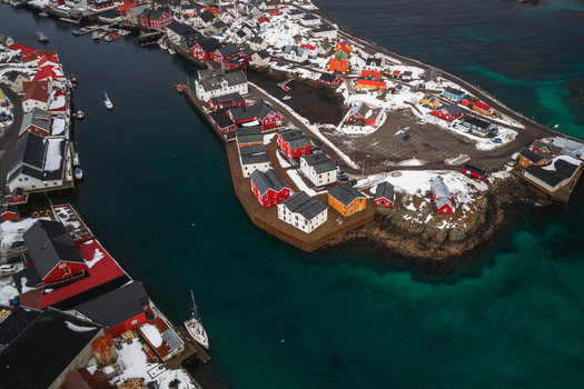 Aerial view of vibrant red and white buildings nestled on rocky shores against the backdrop of dark moody skies and crystal clear turquoise waters Henningsv&aelig;r Nordland Norway.