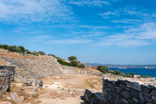 View of the Ancient Theatre of Thorikos with its stone seating tiers overlooking the blue sea and cargo ships under a clear sky in Thoricus Greece.