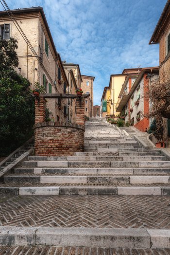View of stone steps leading up through a historic street with a brick well and traditional buildings under a blue sky in Corinaldo Marche Italy. View of stone steps leading up through a historic street with a brick well and traditional buildings under a blue sky in Corinaldo Marche Italy.