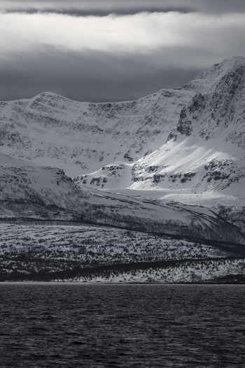 View of dramatic snow covered mountains rising above a dark fjord under a moody cloudy sky in Tromso Troms Norway.