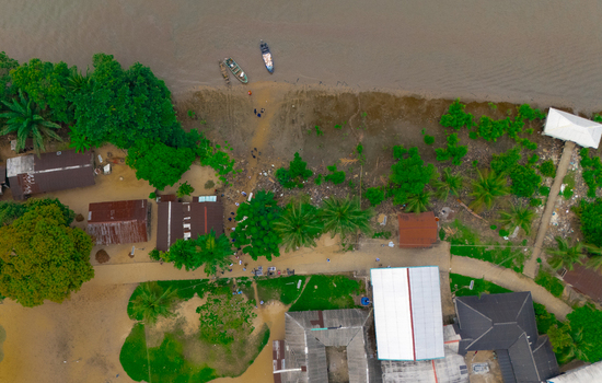 Aerial view of a riverside village with small wooden boats moored at the muddy bank surrounded by lush tropical palm trees and simple houses in Rivers Nigeria.