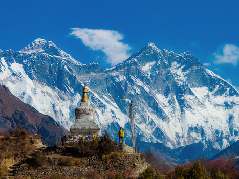 Aerial view of a Buddhist stupa with colorful prayer flags and a man standing on a stone platform against the backdrop of the snow capped Mount Everest peaks in Nepal.