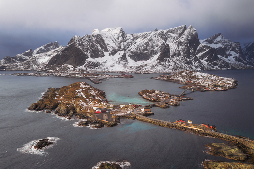 Aerial view of snow capped mountains rising majestically behind the quaint fishing village connected by a bridge Reinebringen Nordland Norway.