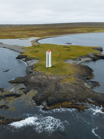 Aerial view of a solitary white lighthouse with a red roof standing proudly on a rocky outcrop amidst the dark blue sea K&aacute;lfshamarsv&iacute;k H&uacute;nabygg&eth; Iceland.