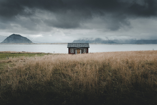 View of a small red wooden house in a field of dry grass with mountains and a fjord in the background under a dark cloudy sky in Nordland Norway. View of a small red wooden house in a field of dry grass with mountains and a fjord in the background under a dark cloudy sky in Nordland Norway.