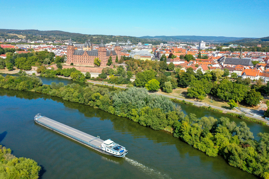 Aerial view of Schloss Johannisburg and a cargo barge navigating the Main river surrounded by lush greenery and the city skyline in Aschaffenburg Bayern Germany.