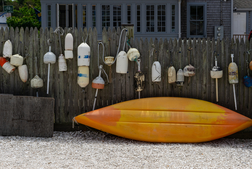 View of a yellow kayak resting on white gravel in front of a wooden fence decorated with weathered fishing buoys in Edgartown Massachusetts United States.