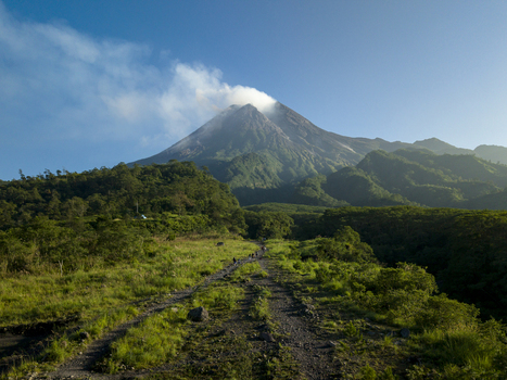 Aerial view of Bunker Kaliadem Merapi and the smoking crater of Mount Merapi volcano with lush green slopes and a dirt path Kecamatan Pakem Daerah Istimewa Yogya Indonesia. Aerial view of Bunker Kaliadem Merapi and the smoking crater of Mount Merapi volcano with lush green slopes and a dirt path Kecamatan Pakem Daerah Istimewa Yogya Indonesia.