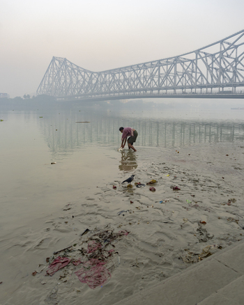 View of a man wading through the muddy riverbank under the majestic Howrah Bridge casting a long shadow on the water in Kolkata West Bengal India. View of a man wading through the muddy riverbank under the majestic Howrah Bridge casting a long shadow on the water in Kolkata West Bengal India.