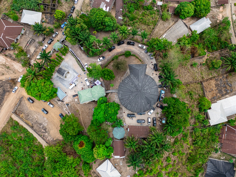 Aerial view of a circular church with a dark roof amidst vibrant green foliage and dusty paths bustling with parked cars Asaga Abia Nigeria. Aerial view of a circular church with a dark roof amidst vibrant green foliage and dusty paths bustling with parked cars Asaga Abia Nigeria.