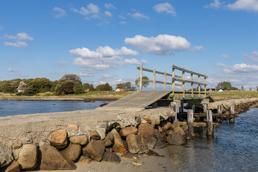 View of a wooden footbridge on a stone causeway extending into the sea on Hjarno with a grassy coastline under a clear blue sky Hjarno Hedensted Municipality Denmark. View of a wooden footbridge on a stone causeway extending into the sea on Hjarno with a grassy coastline under a clear blue sky Hjarno Hedensted Municipality Denmark.