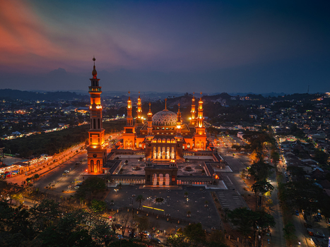 Aerial view of the illuminated Baitul Muttaqien Mosque at the East Kalimantan Islamic Center featuring glowing domes and minarets against a dusk sky in Samarinda East Kalimantan Indonesia.