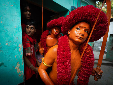 Munshiganj Bangladesh 13 April 2025 View of vibrant orange painted figures with elaborate crimson headwear and garlands emerging from a teal doorway their solemn faces contrast darks and lights. Munshiganj Bangladesh 13 April 2025 View of vibrant orange painted figures with elaborate crimson headwear and garlands emerging from a teal doorway their solemn faces contrast darks and lights.