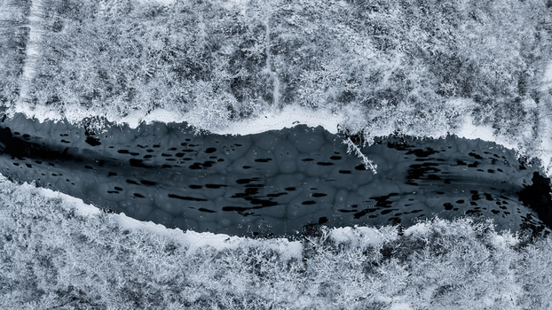 Aerial view of a frozen river with dark patterns on the ice surface surrounded by a dense forest covered in fresh white snow in Latvia. Aerial view of a frozen river with dark patterns on the ice surface surrounded by a dense forest covered in fresh white snow in Latvia.