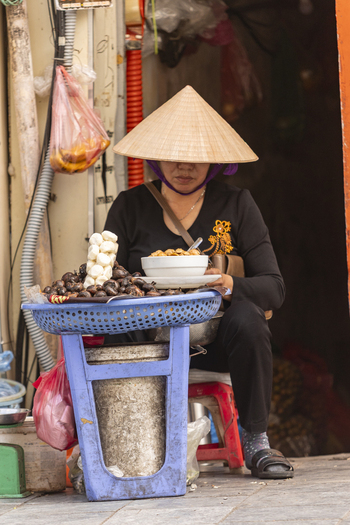 View of a street vendor wearing a traditional conical hat selling snacks from a blue plastic stand on a sidewalk in Hanoi Ha Noi Vietnam.