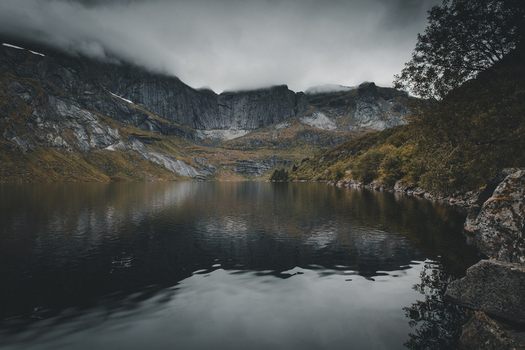 View of the dramatic Batman Wall mountain range reflecting in the dark still waters of a lake under a cloudy sky in Ramberg Nordland Norway. View of the dramatic Batman Wall mountain range reflecting in the dark still waters of a lake under a cloudy sky in Ramberg Nordland Norway.
