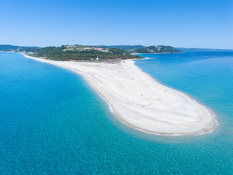 Aerial view of the white sand spit of Possidi Beach surrounded by turquoise waters and lush green trees under a clear blue sky in Greece.