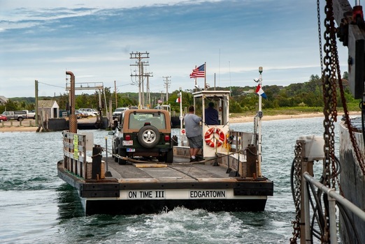 View of the Chappaquiddick ferry On Time III carrying a vehicle across the water with crew members and an American flag Edgartown Massachusetts United States.