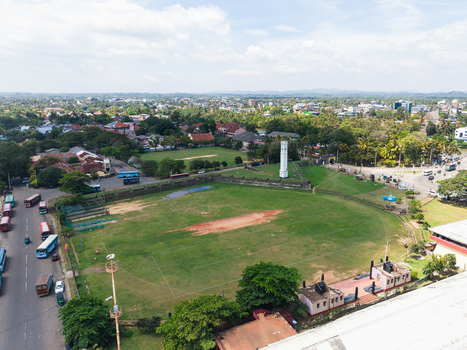 Aerial view of the historic white clock tower and a large green cricket field surrounded by urban buildings and lush trees in Matara Southern Province Sri Lanka. Aerial view of the historic white clock tower and a large green cricket field surrounded by urban buildings and lush trees in Matara Southern Province Sri Lanka.
