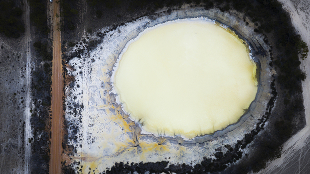 Aerial view of a circular salt lake with pale yellow water surrounded by dry textured terrain and a dirt track in Perth Western Australia Australia.
