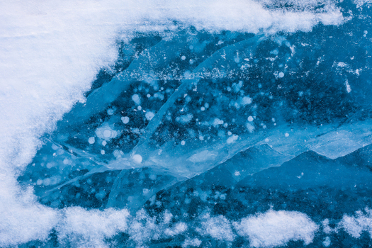 View of Lake Baikal featuring intricate patterns of frozen methane bubbles trapped within the deep blue ice surrounded by patches of white snow in Severobaykalsk Buryatia Russia. View of Lake Baikal featuring intricate patterns of frozen methane bubbles trapped within the deep blue ice surrounded by patches of white snow in Severobaykalsk Buryatia Russia.