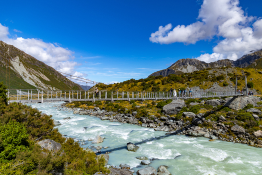 View of a suspension bridge stretching over milky glacial river under a vibrant blue sky connecting rocky terrains Mount Cook Canterbury Region New Zealand. View of a suspension bridge stretching over milky glacial river under a vibrant blue sky connecting rocky terrains Mount Cook Canterbury Region New Zealand.