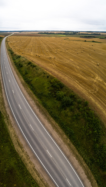 Aerial view of a paved road curving through golden agricultural fields dotted with hay bales under an overcast sky in Latvia. Aerial view of a paved road curving through golden agricultural fields dotted with hay bales under an overcast sky in Latvia.