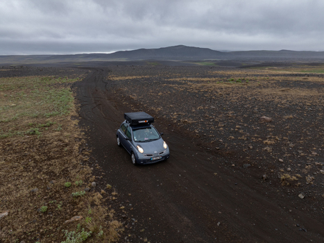 Aerial view of a small car with a roof tent driving along a dark volcanic dirt road through the vast barren landscape of Iceland. Aerial view of a small car with a roof tent driving along a dark volcanic dirt road through the vast barren landscape of Iceland.