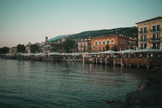 View of Lake Garda with historic waterfront buildings restaurants with white umbrellas and wooden piers under a soft evening sky in Italy. View of Lake Garda with historic waterfront buildings restaurants with white umbrellas and wooden piers under a soft evening sky in Italy.