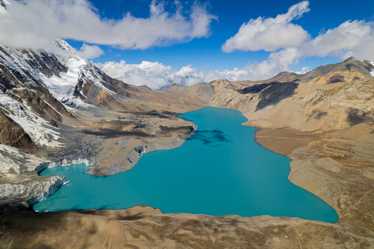 Aerial view of a turquoise glacial lake surrounded by snow capped Himalayan peaks and rugged rocky terrain in Nepal.