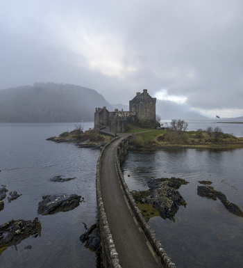 Aerial view of Eilean Donan Castle and its iconic stone bridge surrounded by the calm waters of Loch Duich under a misty sky in Dornie Scotland United Kingdom.