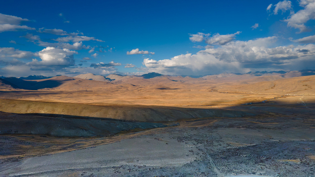 Aerial view of Deosai National Park featuring vast golden plains rolling hills and dramatic cloud formations under a clear blue sky in Skardu Gilgit Pakistan.