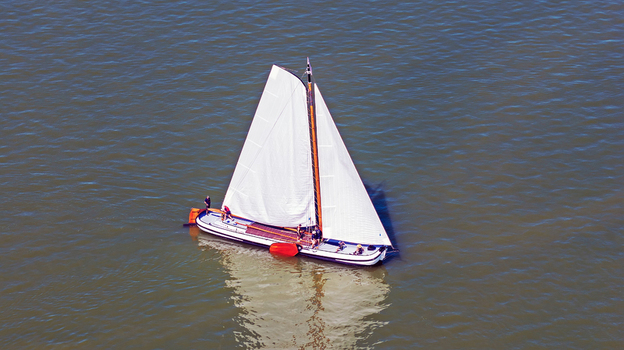 Aerial view of a traditional Dutch sailing boat with white sails gliding across the water with people on deck in Heeg Friesland Netherlands. Aerial view of a traditional Dutch sailing boat with white sails gliding across the water with people on deck in Heeg Friesland Netherlands.
