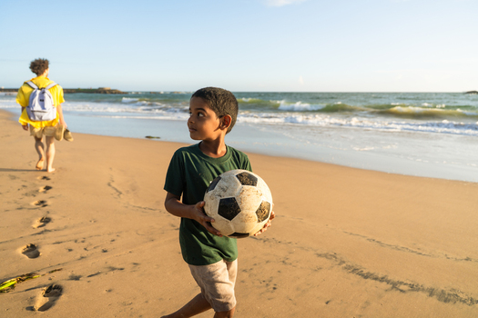 Sri Lanka 11 January 2026 View of a young boy holding a soccer ball on a sandy beach with a man walking in the background during golden hour.