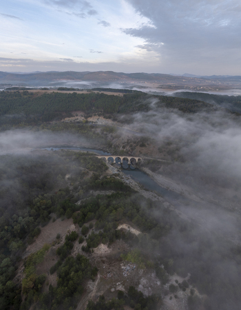 Aerial view of the historic railway bridge spanning a river amidst rolling hills dense forests and low hanging morning mist in Balabanovo Kardzhali Province Bulgaria. Aerial view of the historic railway bridge spanning a river amidst rolling hills dense forests and low hanging morning mist in Balabanovo Kardzhali Province Bulgaria.