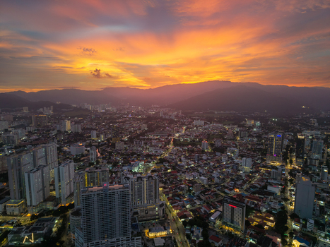George Town Malaysia 22 September 2026 Aerial view of George Town cityscape at sunset with illuminated skyscrapers residential buildings and distant mountains under a vibrant orange sky.