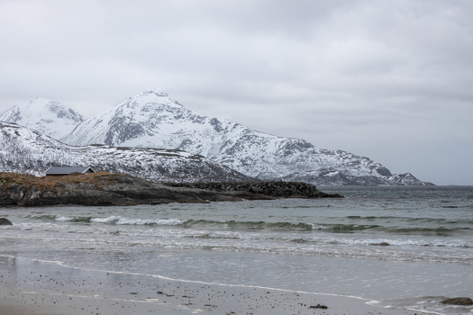View of a sandy beach with gentle waves a small wooden cabin on a rocky shore and majestic snow covered mountains under a cloudy sky in Kvaloya Troms Norway. View of a sandy beach with gentle waves a small wooden cabin on a rocky shore and majestic snow covered mountains under a cloudy sky in Kvaloya Troms Norway.
